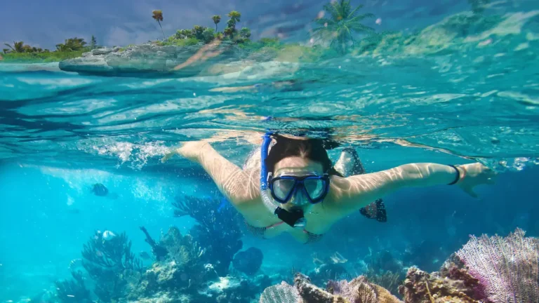 Femme pratiquant le snorkeling avec un masque bi-hublot bleu et tuba, nageant en surface au-dessus de coraux dans une eau turquoise cristalline