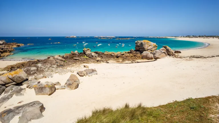 Plage de sable blanc bordée de rochers granitiques avec eau turquoise et bateaux ancrés dans la baie, archipel des Glénan en Bretagne