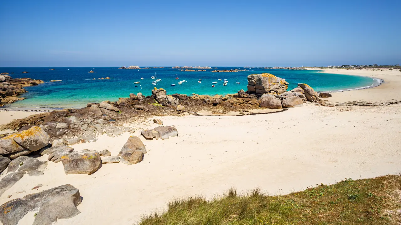 Plage de sable blanc bordée de rochers granitiques avec eau turquoise et bateaux ancrés dans la baie, archipel des Glénan en Bretagne